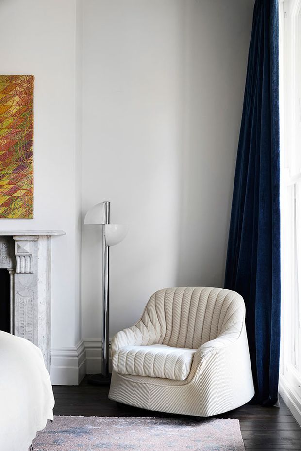 Elegant bedroom corner featuring a sculptural cream upholstered armchair with ribbed detailing, dark timber flooring, white walls with decorative moulding, a classic white fireplace, a tall chrome-and-glass floor lamp, and full-height navy curtains beside a bright window. A portion of the bed and a patterned rug are visible.