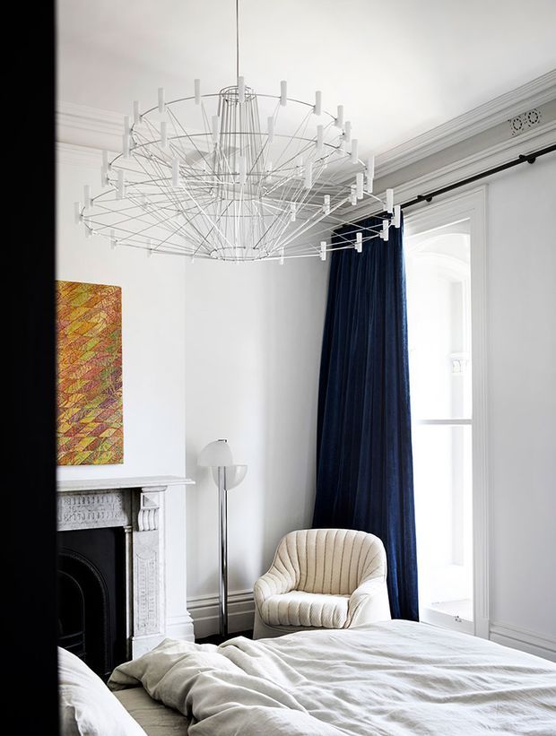 Bright, elegant bedroom with a large sculptural contemporary chandelier, white walls and decorative ceiling cornices, a traditional marble fireplace, tall arched window detailing, deep navy curtains, and a cream upholstered armchair. The room blends classic architectural elements with refined contemporary furnishings.