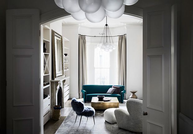 A refined living room viewed through open double doors, with ornate wall panelling and trim, built-in shelving, a fireplace surround, tall curtained window, teal sofa, sculptural cream lounge chairs, a low timber coffee table, and dramatic clustered globe pendant lights. The space has a bright, elegant, contemporary-classic character with strong contrast between the dark entry surround and light interior.