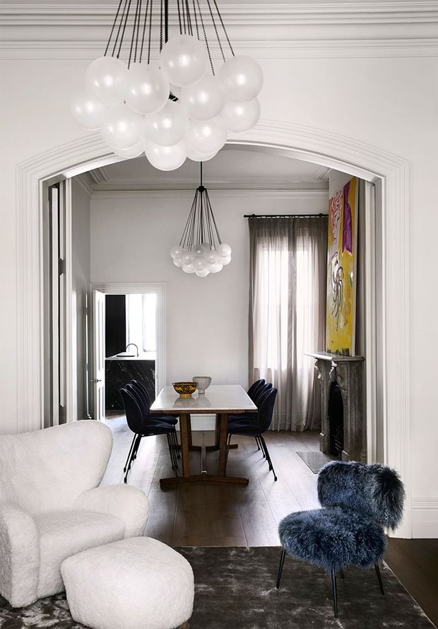 A refined historic-style interior viewed through a wide ornate arched opening, combining a foreground sitting area with a formal dining room beyond. The room features white decorative wall and ceiling mouldings, a dark timber dining table with deep blue chairs, pale timber flooring, a marble or dark stone fireplace, sheer curtains, and striking clustered white globe pendant lights. A colourful artwork adds a bold accent beside the fireplace, while a white boucle lounge chair and dark blue furry accent chair occupy the foreground.