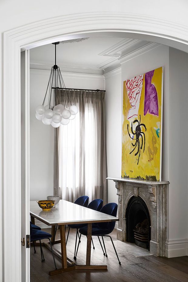 A refined dining room viewed through a wide decorative archway, featuring a long white rectangular dining table with dark blue upholstered chairs, a sculptural cluster pendant light, full-height sheer curtains, ornate ceiling cornices and wall mouldings, and a carved grey stone fireplace. A large colourful abstract artwork hangs above the fireplace. The room combines classic architectural detailing with contemporary furnishings.