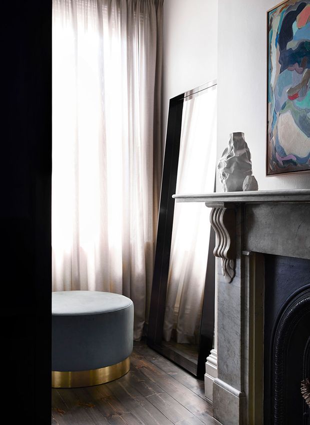 A refined living room corner with tall sheer curtains, a dark wood floor, a round upholstered ottoman with a brass base, a leaning full-length mirror, and an ornate stone fireplace mantel. Abstract artwork and a sculptural vase add visual interest. The room has a sophisticated, layered and softly lit atmosphere.