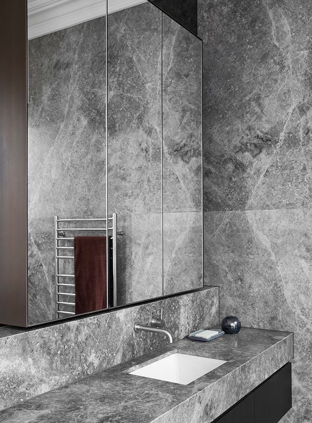 A refined contemporary bathroom vanity clad extensively in grey veined stone, with a large mirrored cabinet, integrated stone backsplash, undermount white basin, wall-mounted chrome tapware, dark timber cabinetry, and a chrome heated towel rail with a deep burgundy towel. The space has a dramatic, luxurious monochromatic appearance with classic ceiling cornice detailing.