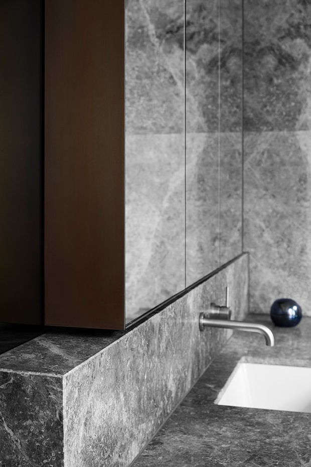Close-up of a refined bathroom vanity featuring dark natural stone benchtops and matching stone wall surfaces, a white inset basin, brushed metal wall-mounted tapware, a dark timber-toned wall cabinet, and a large mirror. The composition is monochromatic and luxurious with strong material contrast between veined stone, timber, and metal.