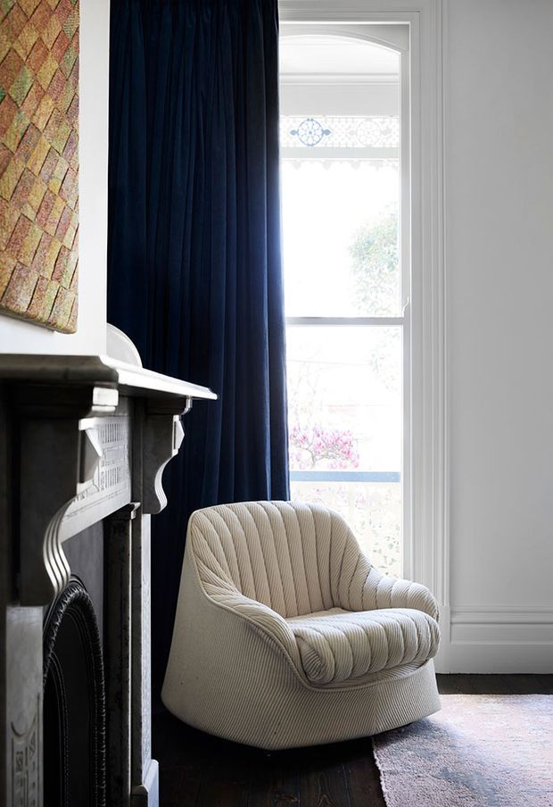 A refined sitting area featuring a sculptural cream upholstered armchair beside a dark traditional fireplace. Deep navy curtains frame a tall decorative window, while white wall mouldings, a patterned artwork, dark timber flooring, and a muted rug create a sophisticated layered interior.