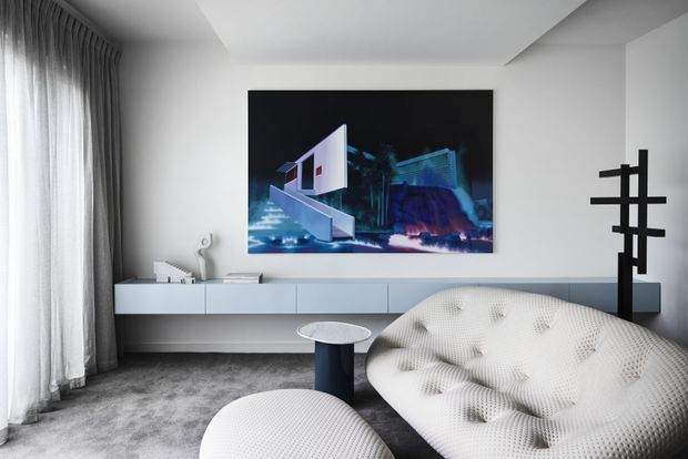 A refined contemporary living room with a low wall-mounted white media cabinet, large abstract artwork, sculptural white tufted lounge seating, a small round side table, dark textured carpet, sheer floor-to-ceiling curtains, and a black geometric floor lamp. The room has a restrained monochromatic palette with strong sculptural forms and soft natural light.