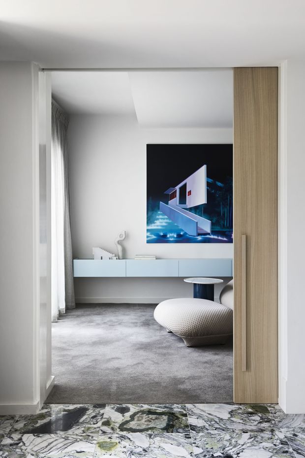 A refined contemporary living room viewed through a wide doorway, featuring a grey carpet, white walls and ceiling, a floating white media console, floor-length curtains, a sculptural low lounge chair, and a small round side table. A large architectural artwork anchors the far wall. The doorway includes a light timber sliding panel, while the foreground hall has a dramatic patterned natural-stone floor.