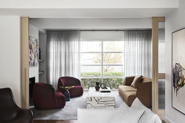 A bright, refined living room with floor-to-ceiling glazing and sheer curtains, a large neutral sectional sofa, sculptural burgundy lounge chairs, a textured brown sofa, a marble or stone coffee table, wall-mounted artwork, and built-in shelving around a dark fireplace opening. The room has a layered contemporary fit-out with strong geometric lines and soft natural light.