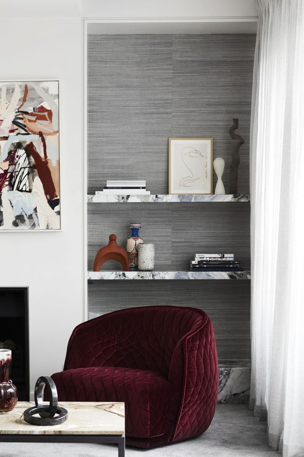 A refined living room vignette featuring a built-in dark grey textured feature wall with open display shelving and pale marble shelves. A deep burgundy quilted lounge chair sits beside a marble-topped coffee table, with sculptural objects, books, artwork, and sheer floor-to-ceiling curtains adding layered detail. The palette is moody and sophisticated, with white walls and soft grey carpet.