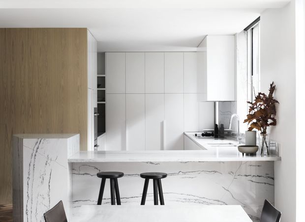 A bright contemporary kitchen with a white marble-look island and waterfall end, integrated white cabinetry, a light timber pantry or wall unit, and black bar stools. The space has clean linear detailing, white walls and ceiling, a stainless-steel sink and tap, and tall side glazing. A small vase with dried foliage provides a warm natural accent.