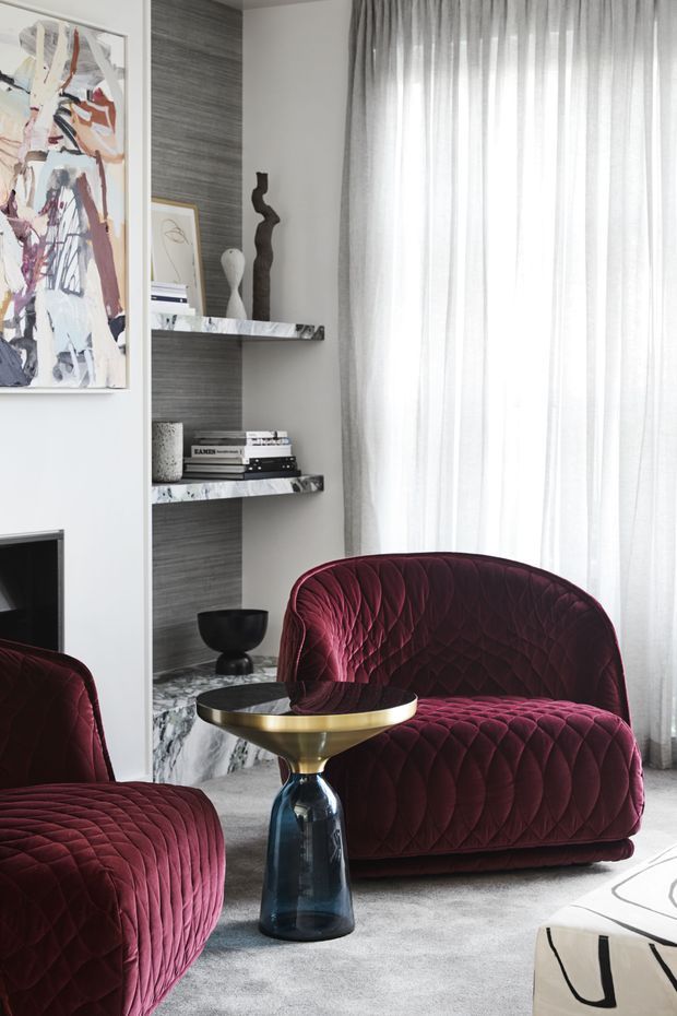 A refined contemporary sitting area featuring deep burgundy quilted lounge chairs, a sculptural dark glass and brass side table, grey carpet, sheer full-height curtains, and a built-in shelving niche with marble shelves and dark textured backing. Abstract artwork and sculptural accessories add visual interest, while the palette is moody and luxurious.
