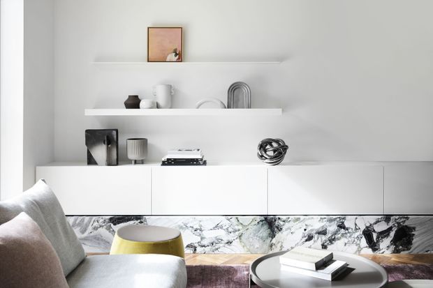 Bright, minimalist living room featuring a long white built-in media or storage cabinet, floating display shelves with curated ceramics and artwork, a pale upholstered sofa, a yellow accent stool, and a round coffee table. A dramatic marble-patterned surface runs beneath the cabinetry, adding contrast to the predominantly white interior.