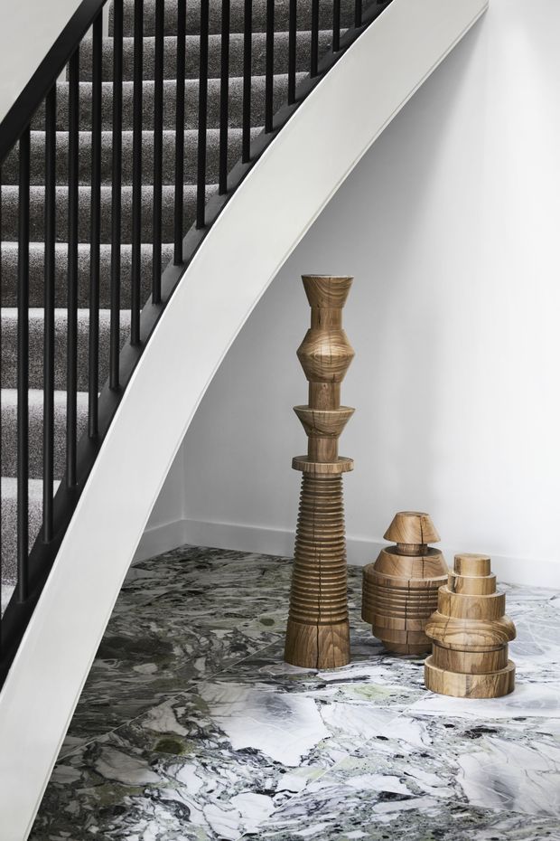 A minimalist stairway with black vertical metal balusters, grey carpeted treads, and a smooth white curved stair stringer. The stair void opens onto a white wall and a dramatic polished grey-and-white marble floor, with sculptural wooden decorative objects arranged beneath the stairs.
