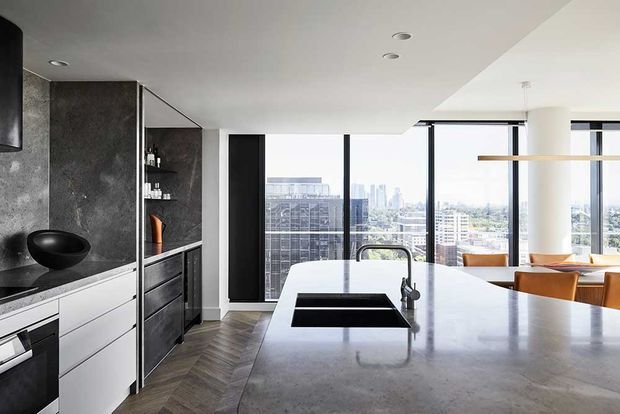 Contemporary high-rise apartment kitchen with a large pale stone island, integrated black double sink and dark tap, dark stone cabinetry and splashback, white lower drawers, and warm timber flooring in a herringbone pattern. Full-height glazing opens to an urban skyline and connects visually to a dining area with timber furniture.