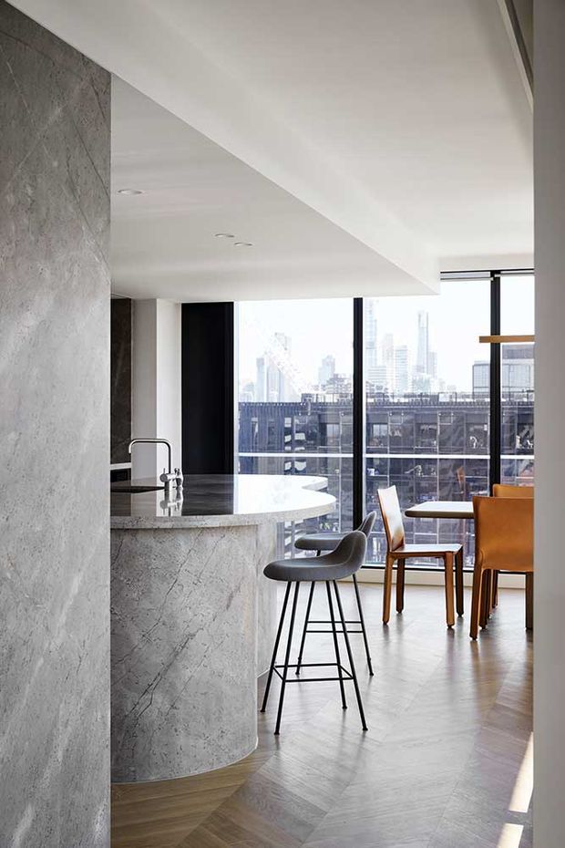 Bright contemporary apartment interior featuring an open kitchen and dining area. The kitchen has a curved island with a grey stone or concrete-look finish, integrated sink and dark tapware, set against a matching textured wall. Floor-to-ceiling black-framed glazing provides expansive city views. A compact dining table and sculptural bar stools sit on a warm herringbone timber floor, creating a refined urban atmosphere.