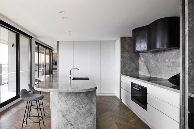 Contemporary kitchen with a large sculptural stone island and integrated sink, white flat-panel cabinetry, dark stone splashback and benchtops, and a distinctive black rangehood. Full-height black-framed glazing lines one side of the room, while herringbone timber flooring adds warmth. Two dark bar stools sit at the island.