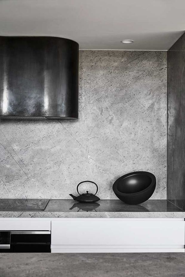 Close-up view of a sleek contemporary kitchen with a dark cylindrical rangehood, grey veined stone backsplash and benchtop, black induction cooktop, black kettle and bowl, and white handleless cabinetry. The composition is monochromatic with dark metal accents and recessed ceiling lighting.