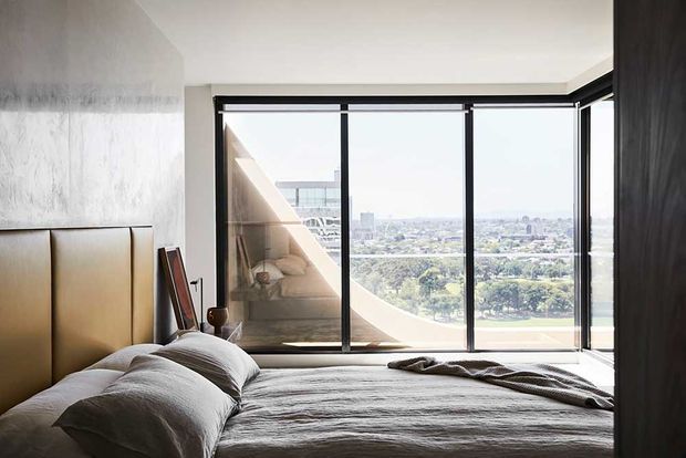 A contemporary bedroom with a large bed, padded tan headboard, textured light wall finish, and expansive black-framed floor-to-ceiling glazing. The window overlooks a leafy urban landscape, while a curved architectural form is reflected in the glass.