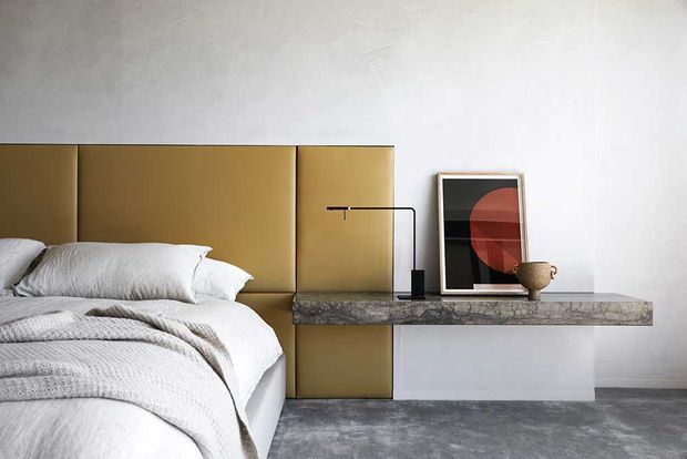 A minimalist bedroom with a low white bed dressed in layered white and grey bedding, backed by a tall mustard-gold upholstered wall panel headboard. A slim floating stone shelf functions as a bedside console, holding a linear black lamp, framed abstract artwork and a ceramic vessel. The room has white plaster walls and a grey textured floor.
