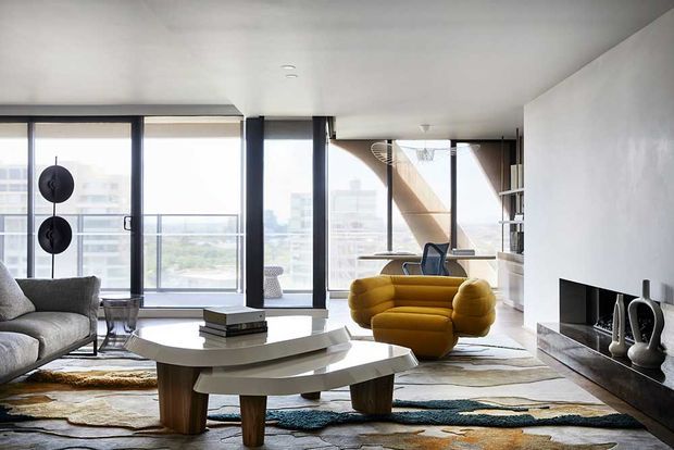 A bright contemporary living room in a high-rise setting, featuring expansive floor-to-ceiling glazing, a grey sofa, a sculptural mustard-yellow lounge chair, layered coffee tables, a linear fireplace set into a dark stone or tile surround, and a large multicoloured area rug. Dark vertical window mullions and a glimpse of built-in shelving reinforce the modern architectural character, with an urban skyline visible beyond the windows.