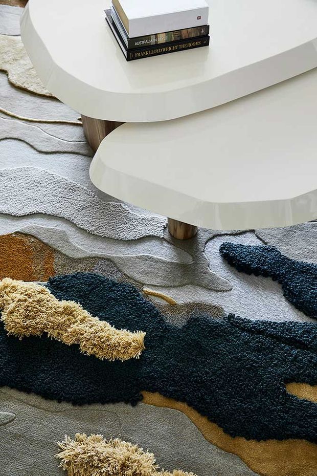 Close-up interior styling detail showing two overlapping white rounded coffee tables with wooden pedestal supports, a stack of books, and a sculptural layered rug in cream, grey, mustard, and deep navy with shaggy textured sections. The composition has a soft, contemporary decorative aesthetic.