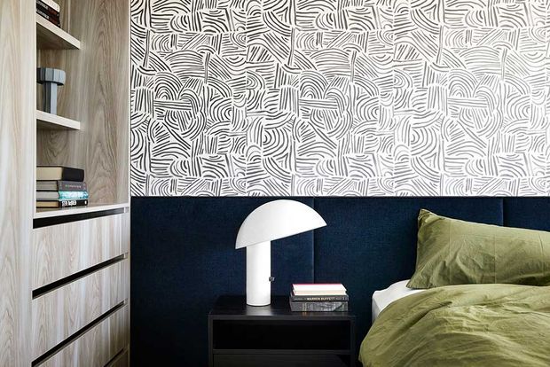 Close-up view of a contemporary bedroom corner featuring a built-in light timber shelving and drawer unit, a deep navy upholstered bedhead, olive-green bedding, and a black bedside table with a white dome-shaped lamp. The wall is covered in a bold black-and-white abstract linear patterned finish.