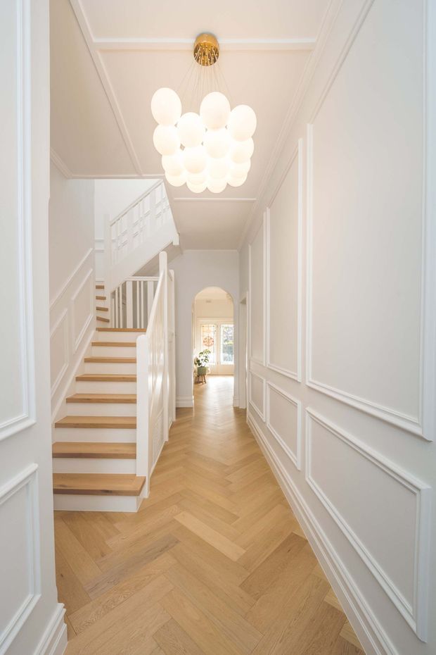 Bright, refined residential hallway viewed from an entry doorway, featuring a warm herringbone timber floor, white decorative wall panelling and moulded ceiling, a white staircase with timber treads, and a prominent clustered spherical pendant light. An arched opening leads toward a sunlit room at the rear.