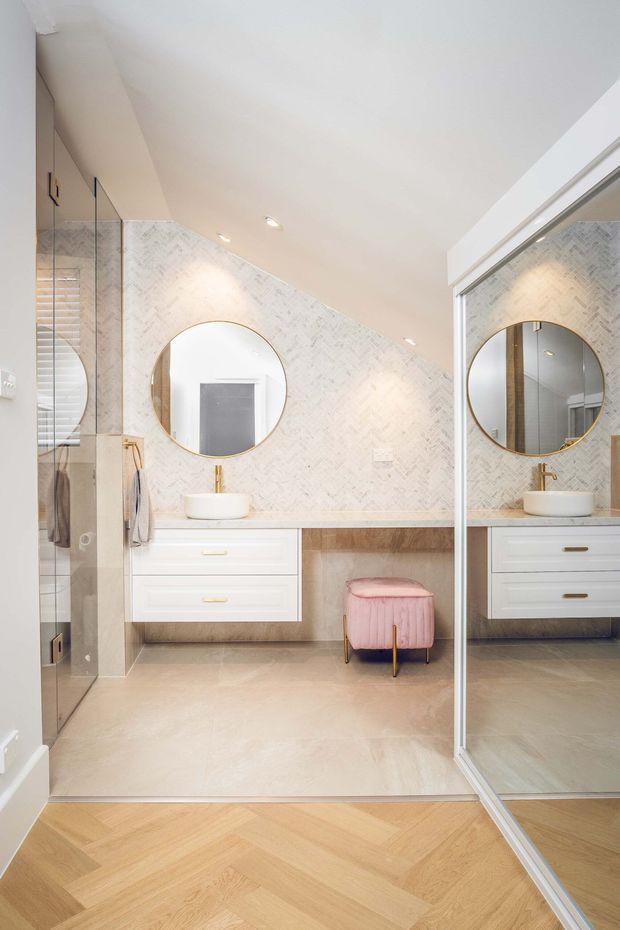 Bright contemporary bathroom with a floating white vanity, vessel basin, brass tapware, round brass-framed mirror, pale herringbone wall tiles, and a glass-enclosed shower. Mirrored wardrobe or cabinetry doors reflect the vanity. A pink upholstered stool adds a soft accent, while warm herringbone timber flooring continues into the foreground.