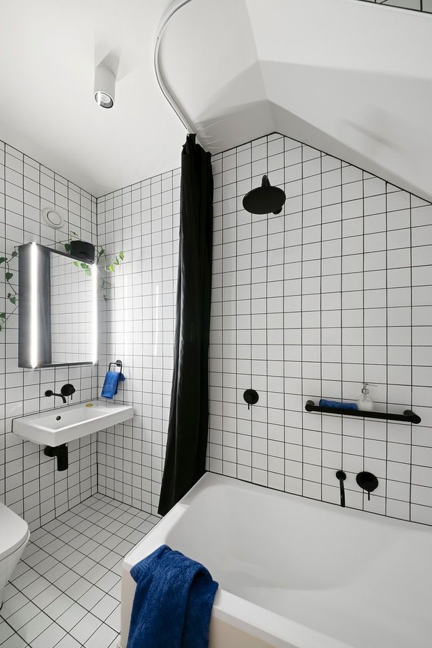 A compact contemporary bathroom with white square grid wall and floor tiles, black grout, matte black shower and tapware, a white freestanding bathtub, wall-mounted white basin, mirrored cabinet, and black shower curtain. Blue towels and small trailing plants provide accents. The room has a bright, graphic, high-contrast aesthetic with an angular sloped ceiling.