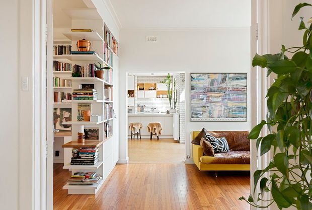 Bright interior view down a timber-floored hallway toward an open kitchen and dining area. Built-in white shelving filled with books and objects lines the left side, while a compact living area with a brown leather sofa and mustard-yellow chair is visible on the right. White walls, wide openings, artwork, and indoor greenery create an airy, eclectic residential interior.