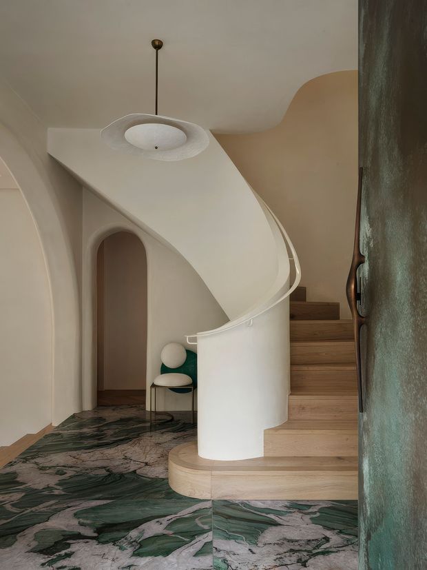A sculptural curved staircase dominates a refined interior, with a white plaster balustrade and organically shaped ceiling canopy. Pale timber stair treads contrast with dramatic green, grey and pink veined stone flooring. An arched doorway, rounded pendant light and a small sculptural chair reinforce the soft, fluid geometry.