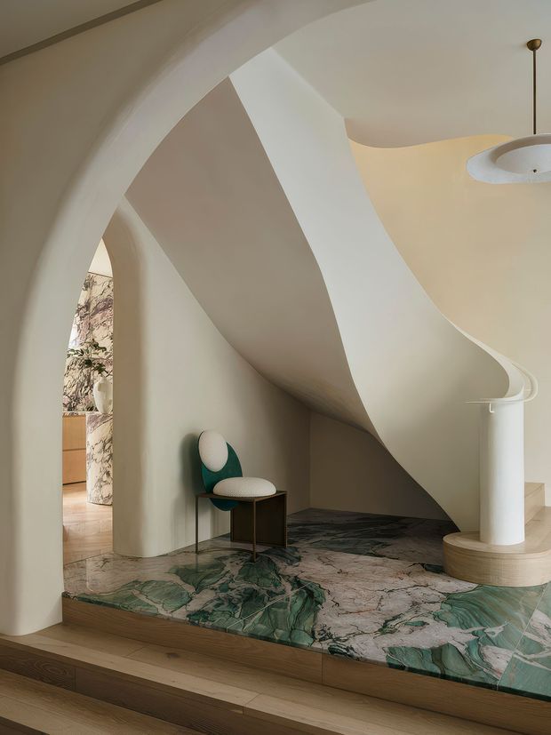 Sculptural contemporary interior featuring flowing, curved plaster arches and a dramatic organic ceiling form. A small sculptural chair sits beside a slender side table on a richly veined green and cream stone platform, with timber steps and a suspended circular light fixture. The space feels gallery-like, refined, and highly architectural.