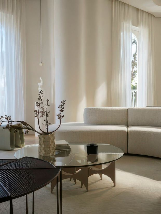 A softly lit contemporary living room with tall sheer-curtained windows, a large curved light upholstered sectional sofa, and sculptural glass coffee tables with pale geometric bases. Neutral beige and cream tones create an airy, refined setting, complemented by sparse botanical branches and dark accent furniture.