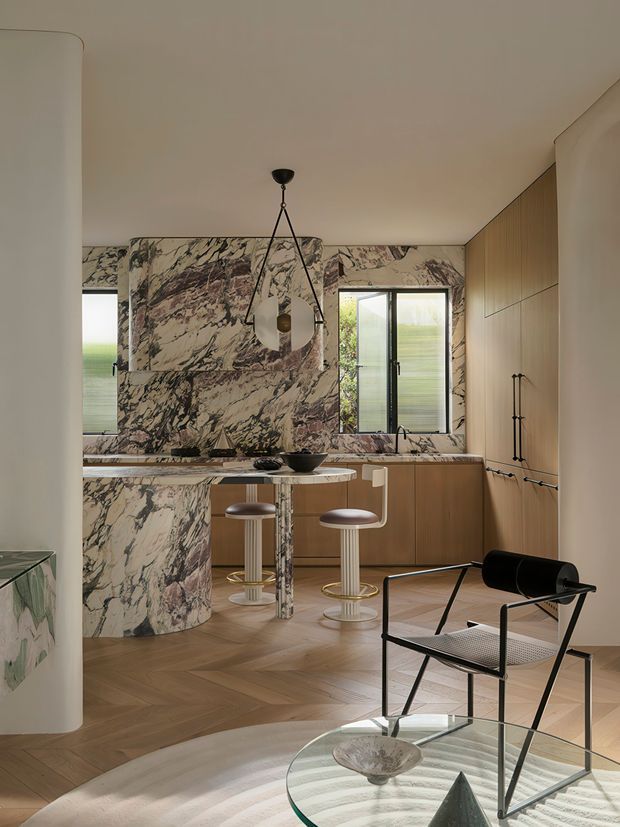 Refined contemporary kitchen and dining space featuring dramatic veined marble wrapping the backsplash, island and counter surfaces, warm timber cabinetry, herringbone timber flooring, and a sculptural hanging pendant. Two bar stools sit at the rounded marble island, while a black-framed lounge chair and glass coffee table occupy the foreground. The space is softly lit and combines luxurious natural stone with warm wood and modern furnishings.
