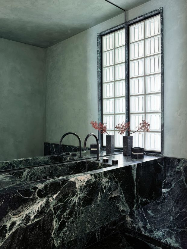 A dramatic, moody bathroom featuring a large sculptural bathtub or integrated bath surround clad in highly veined dark marble. Matching dark marble extends across the vanity or ledge, with two black arched faucets and small dark vessels. Tall multi-pane windows with dark stone or marble surrounds provide soft diffused light, while the walls and ceiling have a muted grey-green plaster finish.