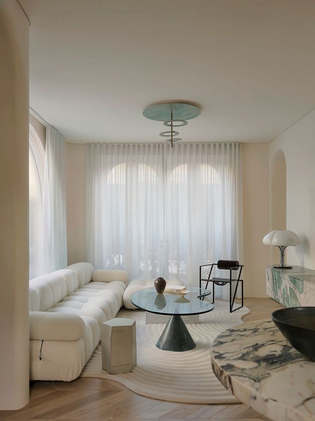A softly lit contemporary living room with sculptural cream seating, a glass coffee table, a black-and-glass side chair, sheer floor-to-ceiling curtains, and a distinctive modern ceiling pendant. Pale walls, arched window forms, light herringbone timber flooring, a textured circular rug, and marble-topped cabinetry create a refined, warm minimalist interior.