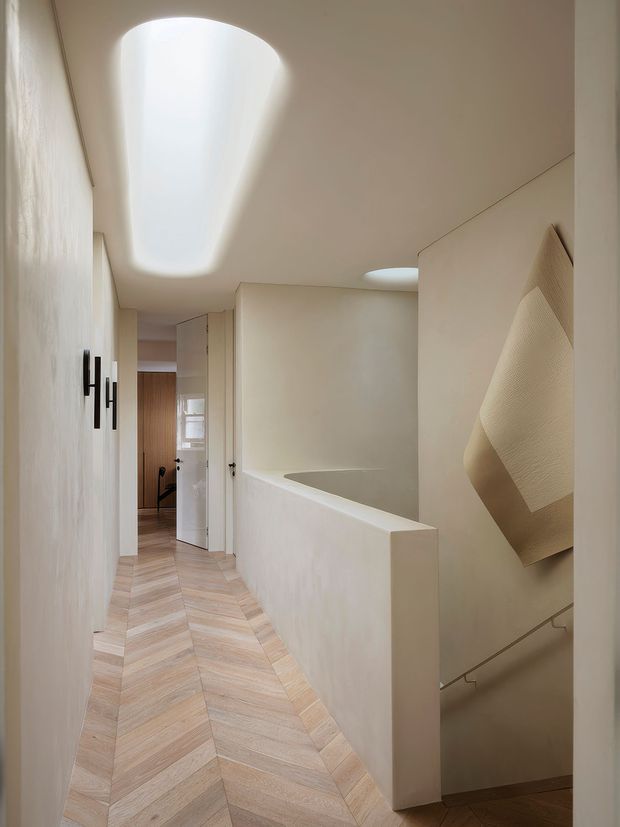 A softly lit minimalist interior hallway and stair landing with curved plaster walls, a sculptural curved skylight, pale herringbone timber flooring, concealed doors, and a partially visible stairwell with an abstract textured wall feature.
