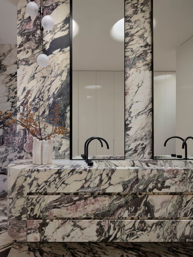 A dramatic, highly patterned bathroom vanity featuring extensive bookmatched-looking natural stone across the countertop, backsplash, drawer fronts, and surrounding walls. Two large mirrored panels reflect the room, while matte black curved faucets and minimal black hardware contrast with the pale marble and dark veining. Globe pendant lights hang at the left, and a sculptural white vase with branches sits on the vanity. The overall composition is luxurious, symmetrical, and richly veined.