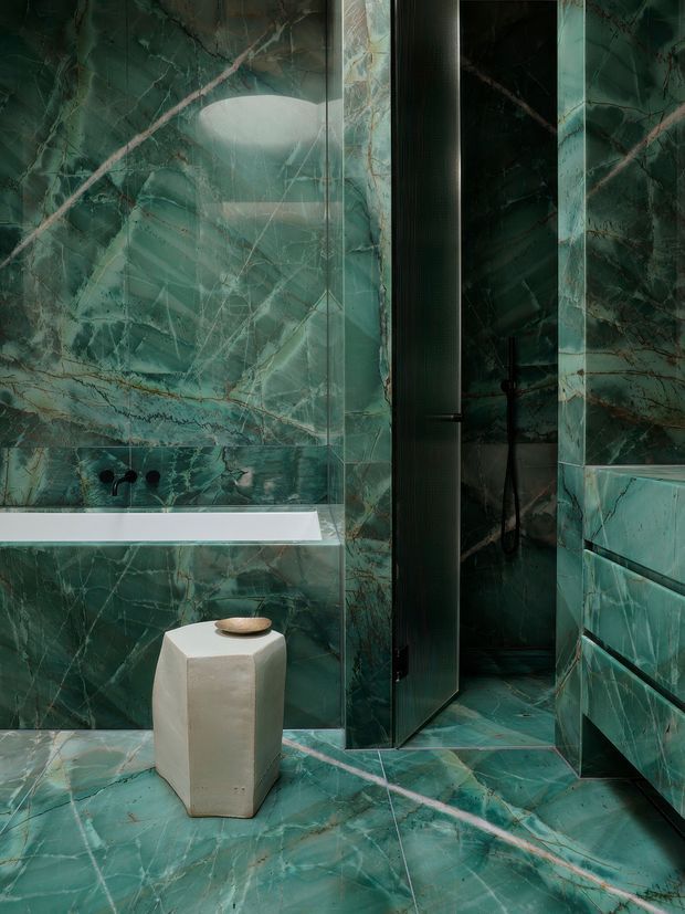 A dramatic, compact bathroom clad almost entirely in richly veined deep green marble or marble-look porcelain slabs. The space includes a wall-mounted bathtub with dark fixtures, a partially open dark shower or bathroom door, a built-in vanity with integrated drawers, and a pale sculptural stool or side table. The dark green surfaces, black hardware, and limited lighting create a luxurious, moody atmosphere.