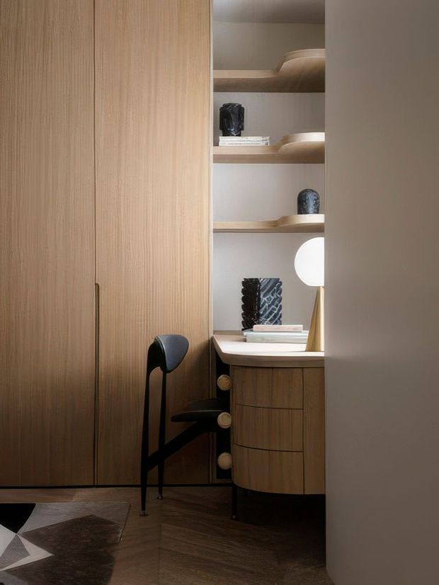 A compact, sophisticated built-in home office or dressing nook with full-height light timber cabinetry, open display shelving, a curved timber drawer unit and pale benchtop. A black sculptural chair sits at the desk, with a round wall light and carefully styled objects adding contrast. Dark herringbone timber flooring and warm, low lighting create an intimate, refined atmosphere.