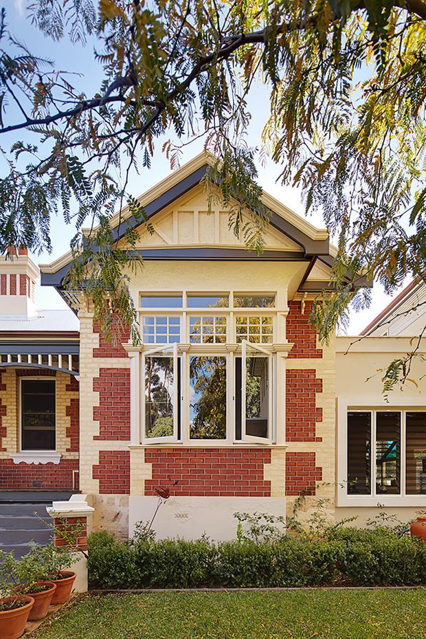 Front exterior view of a heritage-style brick residence, framed by mature tree branches. The facade features red brick with contrasting light rendered bands, a prominent gabled bay, decorative timber gable detailing, cream-trimmed multi-pane windows, and a neatly landscaped front garden. A contemporary or later side extension is visible to the right.