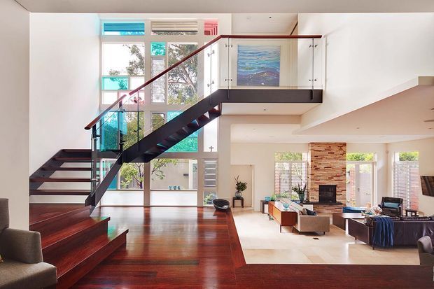 Bright open-plan contemporary interior featuring a dramatic dark timber staircase with glass balustrades, polished dark timber flooring, and a mezzanine level with white built-in cabinetry. Large windows provide abundant natural light, while the lower level opens into a living area with a stone fireplace and exposed brick feature wall.