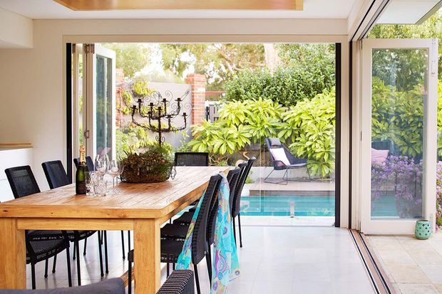 Bright dining area with a large timber dining table and black chairs, opening through extensive sliding and hinged glazing to a landscaped courtyard and swimming pool. The room has pale walls, a light tiled floor, and a timber-lined recessed ceiling feature.