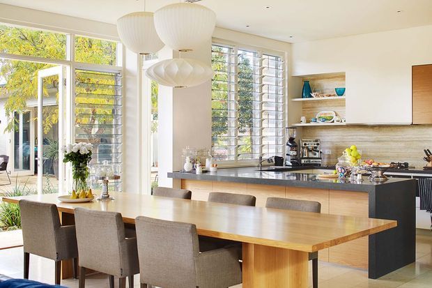Bright open-plan kitchen and dining area with a large timber dining table, upholstered dining chairs, streamlined timber cabinetry, dark island cabinetry, and a stone benchtop. Full-height glazing with horizontal louvres opens to a leafy outdoor setting, while sculptural white pendant lights hang above the dining table.
