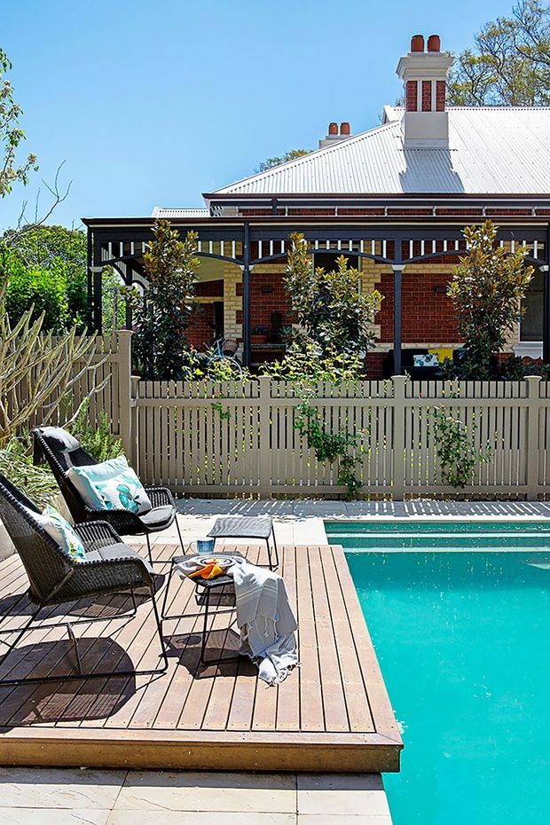 A sunny backyard pool area with a timber deck, turquoise swimming pool, black woven lounge chairs, and a small outdoor table. A pale picket fence and lush planting separate the pool from a traditional red-brick house with a corrugated metal roof, decorative veranda, and prominent chimneys.