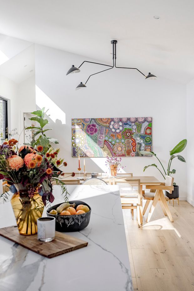 A bright, airy open-plan dining and kitchen interior. A white marble-look island dominates the foreground, styled with a large floral arrangement, fruit bowl and timber serving board. Behind it is a light timber dining table with woven cane chairs, indoor plants, colourful Aboriginal-style artwork, and a sculptural black multi-arm pendant light. White walls, pale timber flooring and strong natural daylight create a warm contemporary setting.
