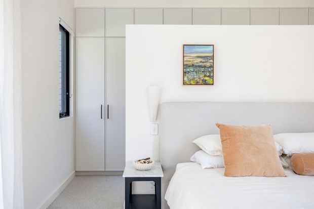 Bright, compact contemporary bedroom with a soft upholstered bedhead, white bedding, pale grey built-in wardrobe cabinetry, and a small black bedside table. A narrow black-framed window, simple wall-mounted light, and restrained artwork contribute to a clean, calm interior.