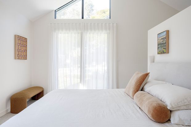 Bright, softly styled bedroom with a white bed, upholstered headboard, layered white bedding, and a long tan bolster cushion. Sheer curtains cover full-height glazing, with an additional high triangular clerestory window bringing in natural light. The room has light neutral walls, minimal furnishings, a small woven stool, and restrained artwork, creating an airy contemporary feel.