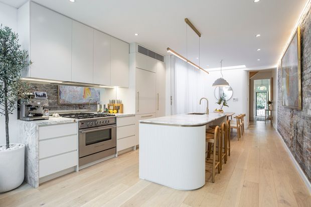 Bright, elongated open-plan kitchen and dining space with a large curved island, white flat-panel cabinetry, integrated appliances, timber flooring, and a stone feature wall. A dining table extends behind the island toward a glazed doorway, while linear and pendant lighting emphasize the contemporary layout.