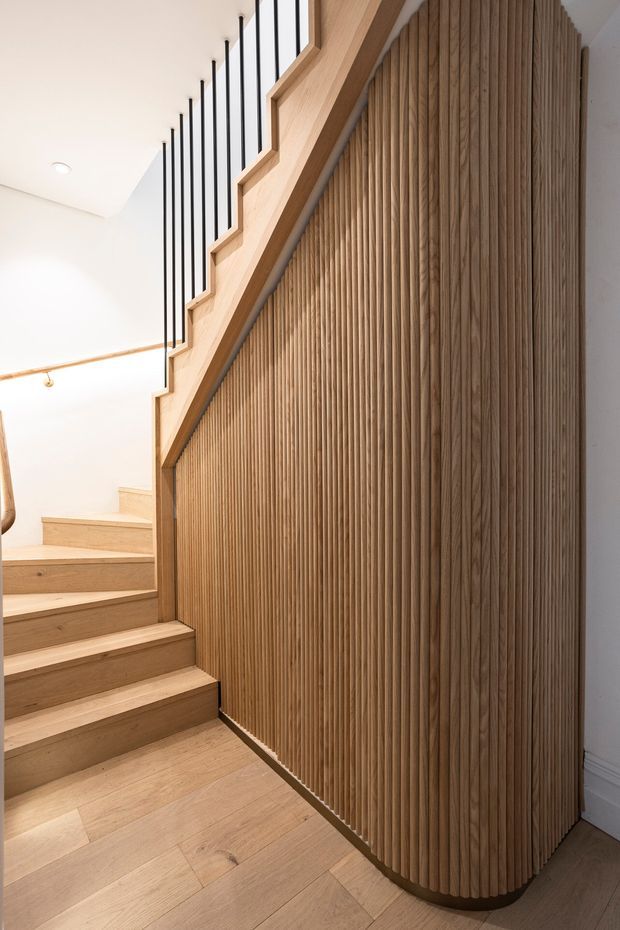 Interior view of a stairway with pale timber flooring and stair treads, a sculptural curved wall clad in narrow vertical timber battens, and a black metal balustrade following the stair profile. White walls and ceiling create a bright, refined, warm-toned space.